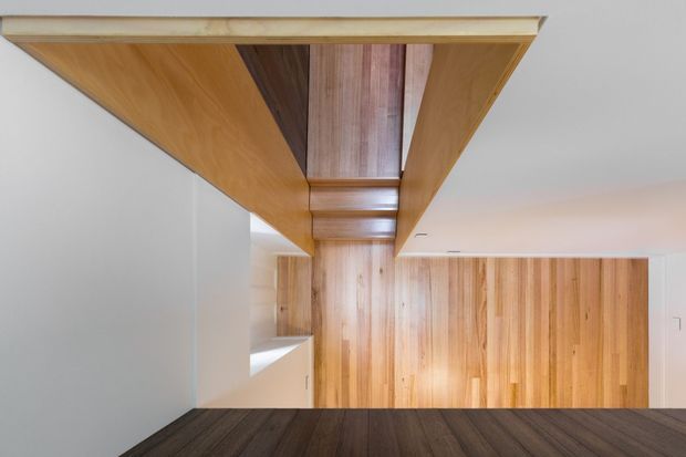 A clean, modern interior hallway or circulation space featuring extensive warm timber lining on the ceiling, upper wall, and doorway, contrasted with smooth white walls and a dark timber floor. The corridor has strong linear geometry, recessed lighting, and integrated timber cabinetry or wall panels.