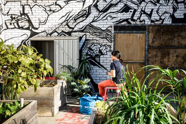 A sunny urban courtyard or rooftop garden with raised timber planter boxes, dense leafy planting, a seated person, and a small corrugated metal shed or storage enclosure. The surrounding walls feature bold black-and-white abstract graffiti-style artwork, with a woven or timber screen on the right. The space has a creative, community-garden atmosphere.