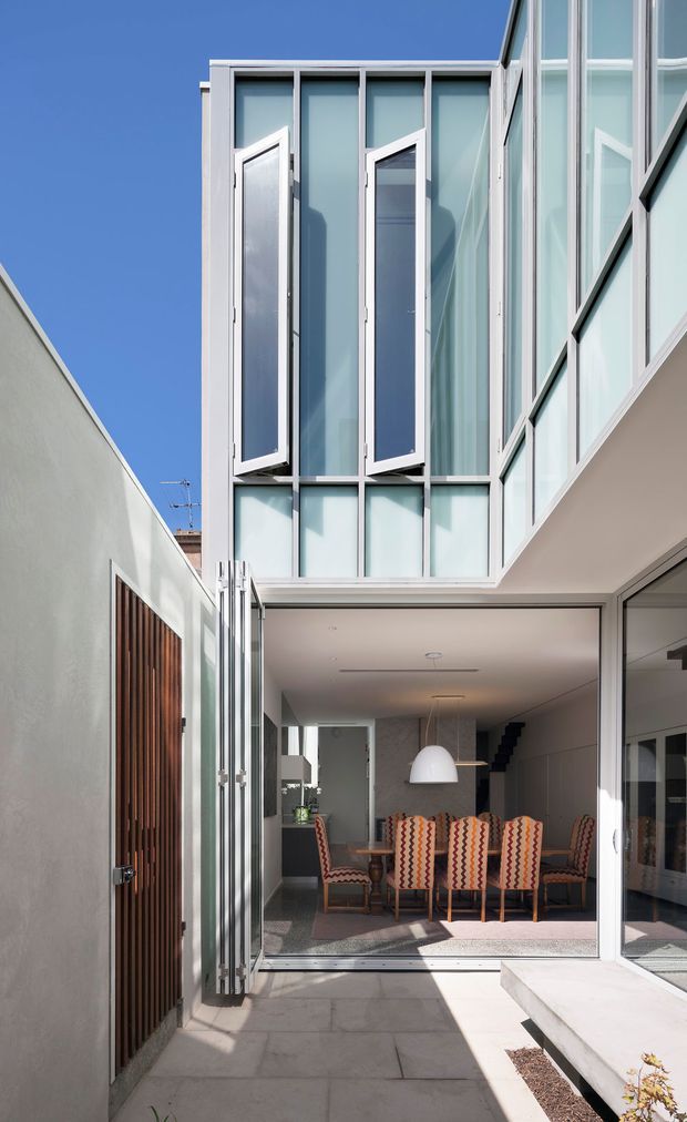 A narrow contemporary residential courtyard or entry passage leading into a dining area. The two-storey extension features extensive frosted and clear glazing in pale aluminium frames, tall opening windows, and a large glazed opening at ground level. Inside, a timber dining table is surrounded by patterned upholstered chairs beneath a white pendant light. A vertical timber slat door and pale rendered walls contrast with the highly glazed modern addition.