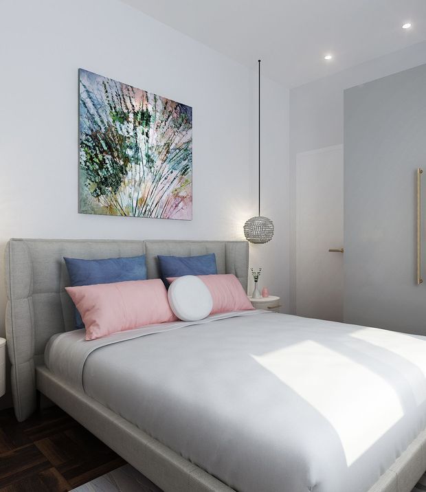 A bright, compact bedroom with a grey upholstered bed and headboard, white bedding, pink and blue accent pillows, and a large botanical abstract artwork above the bed. A spherical pendant light hangs beside the bed, with a small round bedside table and minimal decor. White walls, recessed ceiling lights, dark timber flooring, and a simple white door create a clean contemporary setting.