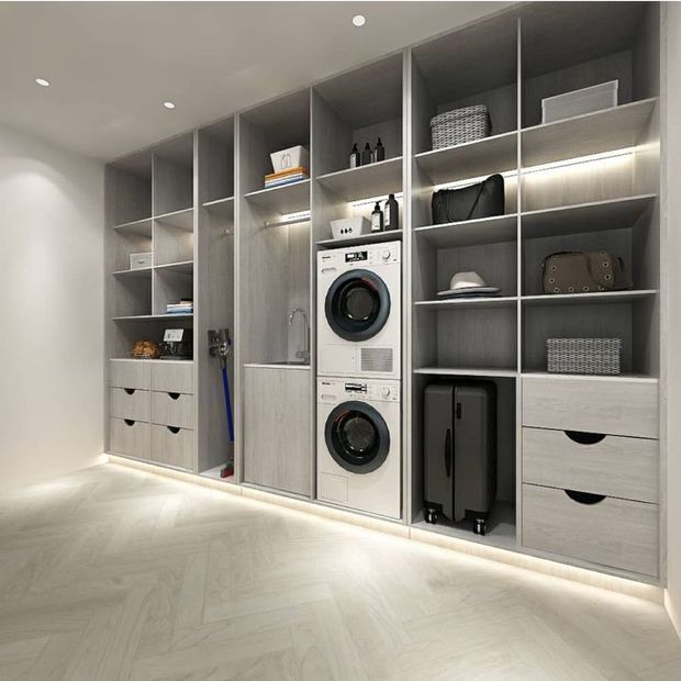 A modern, custom-built laundry and storage wall with stacked front-loading washing and drying machines, an integrated utility sink, open shelving, drawers, hanging space, and concealed LED strip lighting. The space has pale grey timber-look cabinetry, light flooring, and a clean minimalist finish.
