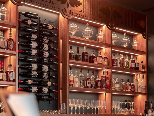 A richly finished built-in bar and wine display with dark timber cabinetry, illuminated open shelving, decorative carved detailing, horizontal wine bottle storage, spirits, decanters, and rows of glassware. The warm integrated lighting creates an intimate, luxurious atmosphere.