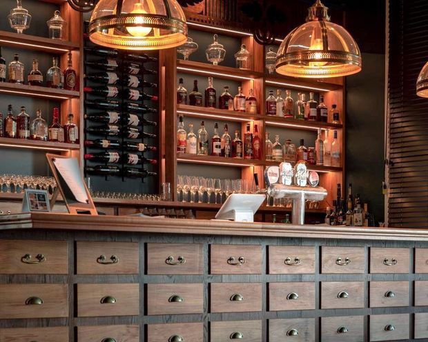A richly detailed bar interior with a long timber counter, extensive built-in bottle shelving, a central wine rack, rows of glassware, beer taps, and warm dome pendant lights. The cabinetry features many small timber drawers with dark metal pulls, creating a refined, atmospheric hospitality setting.