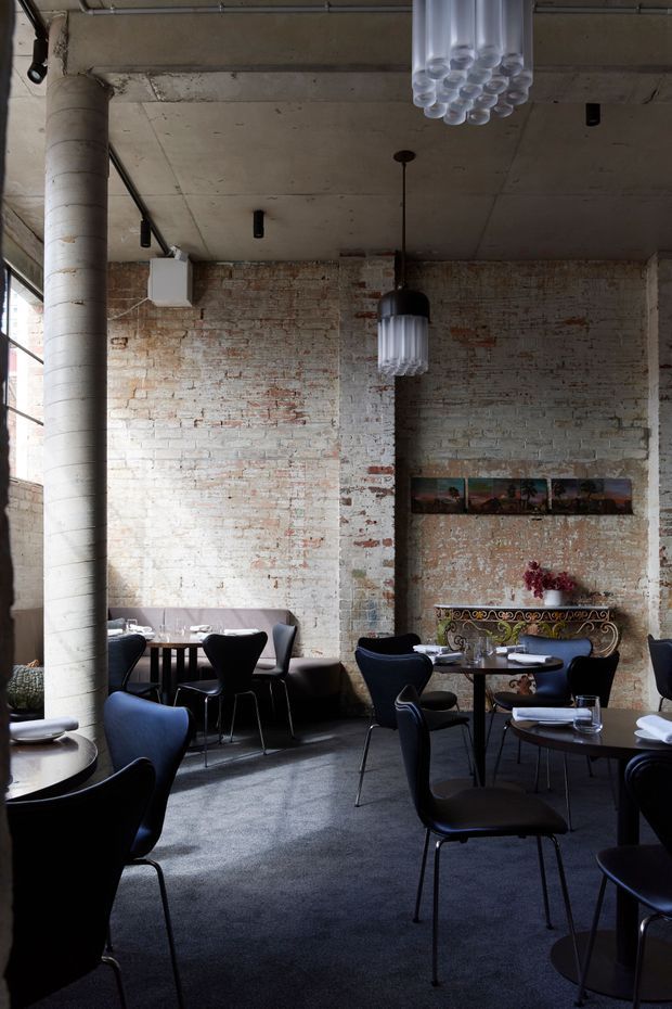 A sophisticated restaurant or hospitality dining interior with exposed whitewashed brick walls, a raw concrete ceiling, a large cylindrical concrete column, and tall industrial-style windows. The room contains multiple small dining tables with dark upholstered chairs, sculptural pendant lights, and an ornate decorative console against the rear wall. The atmosphere is moody, refined, and industrial with eclectic decorative details.
