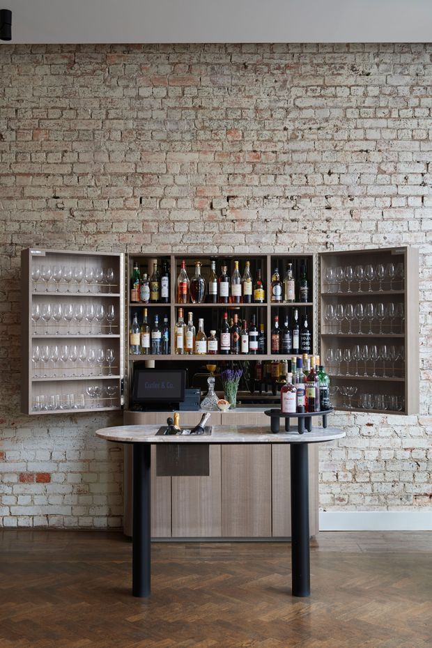 A stylish built-in home bar against an exposed, whitewashed brick wall. The dark timber and glass-backed shelving is stocked with wine and spirits, with extensive glassware storage on both sides. A stone-topped bar counter with black supports and integrated cabinetry stands in front, set on a dark herringbone timber floor. The space has a refined industrial character with warm, muted tones.