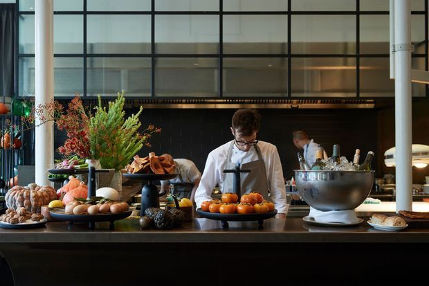 A sophisticated commercial culinary counter or restaurant kitchen viewed from the customer side. Chefs prepare food behind a dark stone counter, with abundant produce and plated items in the foreground. The space features a dramatic black tiled or dark wall backdrop, large black-framed industrial-style glazed panels, cylindrical structural columns, and warm integrated lighting. The composition has a refined, moody hospitality atmosphere.