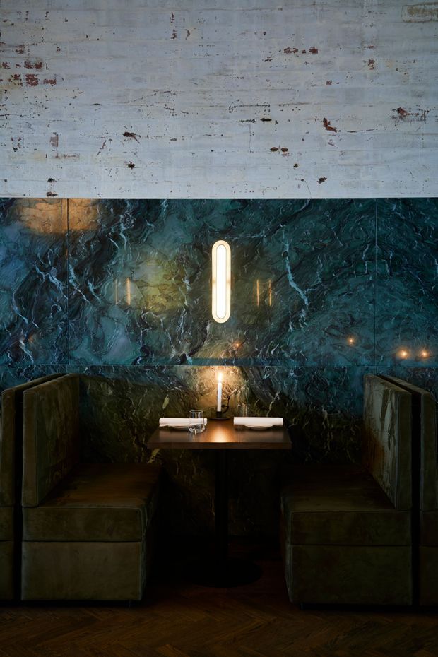 A dark, intimate restaurant booth with two upholstered banquettes facing a small dining table. The walls are clad in dramatic deep teal-green veined stone or marble, contrasted by a weathered pale painted panel above. A slim vertical wall light and candle provide warm illumination, while the dark timber parquet floor reinforces the moody, luxurious atmosphere.
