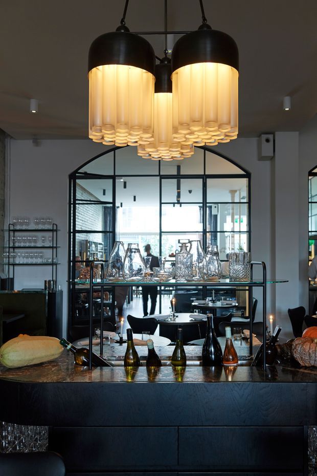 A moody, sophisticated restaurant or hospitality interior centred on a dark bar or service counter. A dramatic cluster of black pendant lights with warm cylindrical shades hangs above a reflective stone counter displaying wine bottles, glassware, decanters and bread. Black-framed glass partitions and an arched glazed opening reveal a dining area beyond, with open shelving and additional tables. The space combines dark joinery, industrial-style glazing and warm ambient lighting.