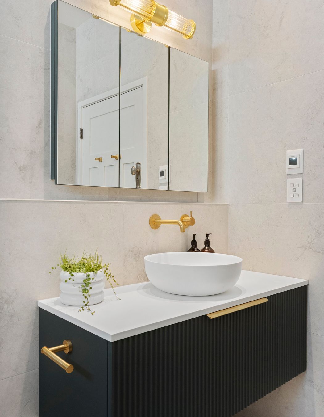 The Vista mirrored wall unit enhances the bathroom's functionality by providing additional storage and convenient power outlets, all while contributing to the sleek design. Its reflective surface amplifies natural light, creating a sense of spaciousness. Complementing this piece, the fluted glass wall lamp perfectly aligns with the fluted vanity and other brushed brass elements, creating a cohesive and harmonious aesthetic. Together, these features add both practicality and style, elevating the overall elegance of the bathroom.