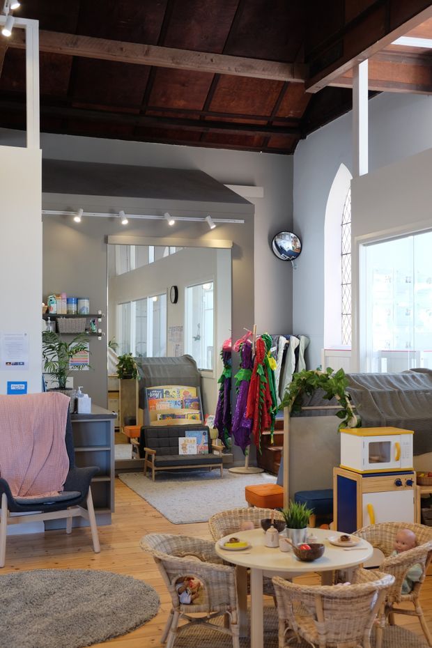 A bright, eclectic multi-purpose interior with a high exposed timber ceiling and beams, pale grey walls, timber flooring, large windows, and a tall arched window. The foreground contains a small children's dining/play area with wicker chairs, a toy kitchen, and a round table, while the background includes lounge seating, shelving, plants, a clothing rack, and a large wall mirror. Track lighting and mixed furnishings create a colourful, informal community or family-oriented space.