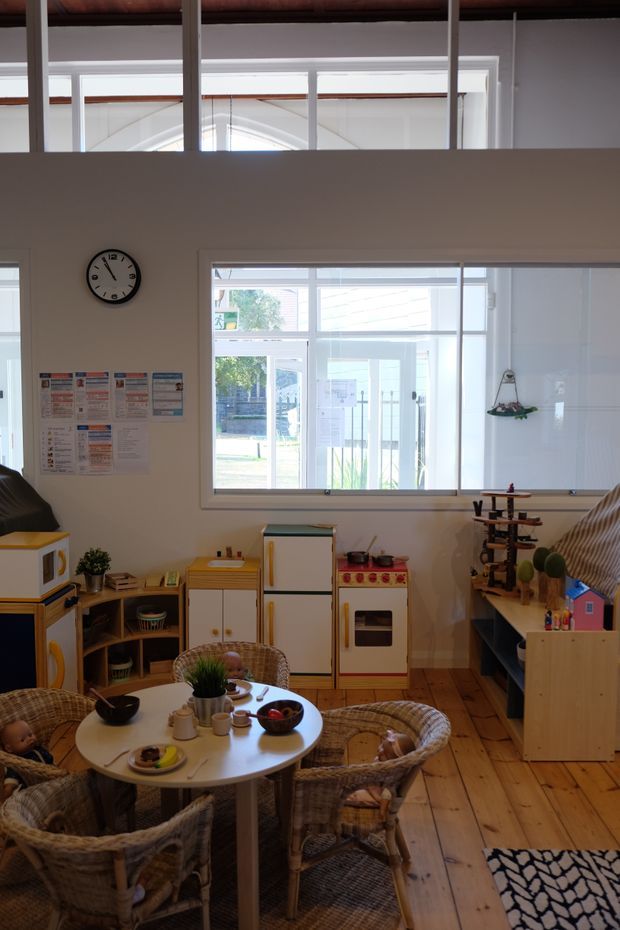 Bright, airy children's playroom or kids' dining area with a small round table surrounded by woven wicker chairs, toy kitchen units, open shelving, and children's toys. The room has white walls, extensive internal glazing and clerestory-style windows, warm timber plank flooring, and exposed dark timber ceiling beams. A large window looks into an adjacent space or exterior area. The overall atmosphere is playful and welcoming, with natural materials and abundant daylight.