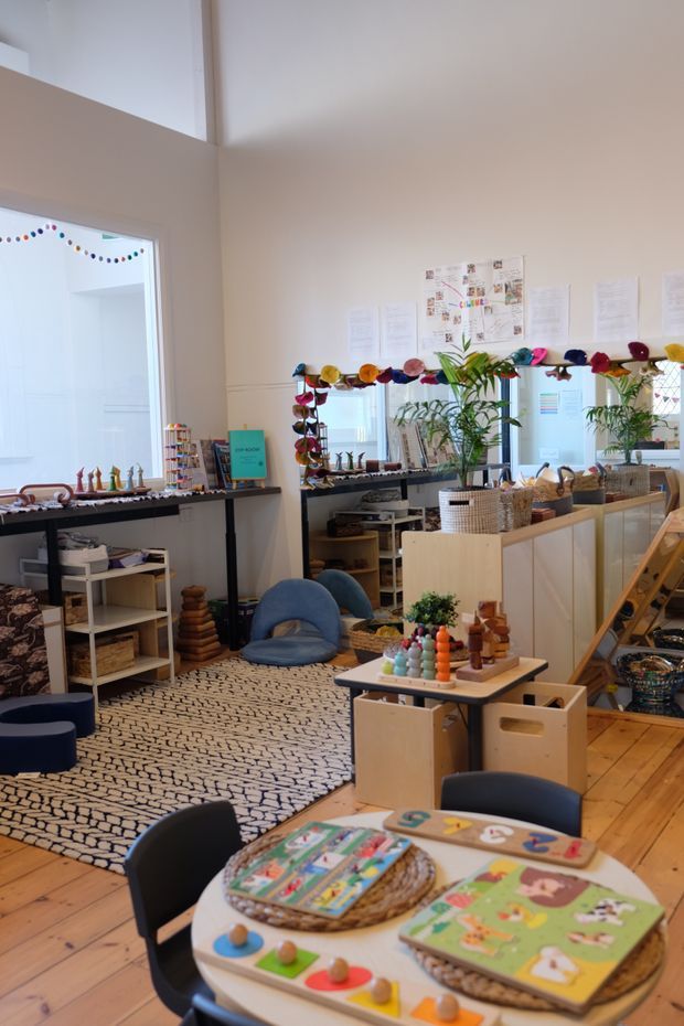 Bright, playful interior of a children's learning or play space, furnished with low activity tables, child-sized chairs, open shelving, educational toys, baskets, plants, and colourful garland decorations. The room has pale walls, timber flooring, a large patterned rug, mirrors, and high ceilings with abundant natural light.