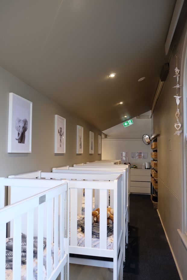 A narrow nursery or childcare room lined with several white baby cots. The space has warm grey walls, a dark carpeted walkway, recessed ceiling lights, framed animal artwork, built-in storage and shelving, and an emergency exit sign at the far end. White cot frames and soft patterned bedding dominate the foreground.