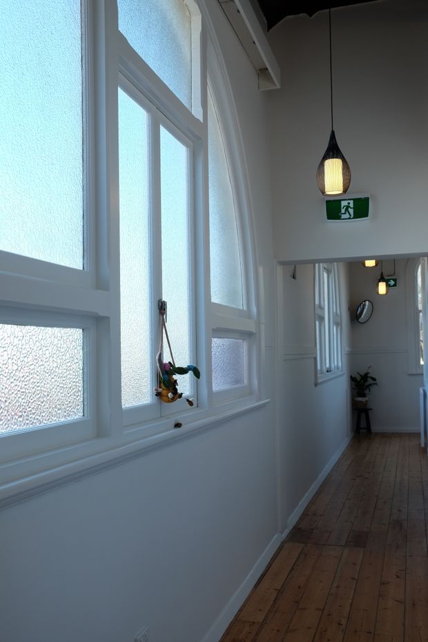 A narrow, bright interior hallway with white-painted walls and trim, a series of frosted glass windows in white frames, and warm natural timber plank flooring. A suspended woven pendant light and green emergency exit signs add visual detail, while a mirror and potted plant are visible farther down the corridor.