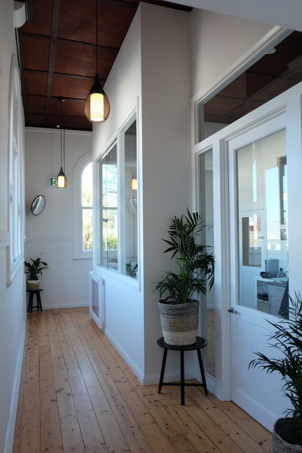 A bright, narrow interior hallway with white walls, framed glazed doors and internal windows, a warm timber plank floor, and a dark timber-panelled ceiling. Pendant lights, decorative wall moulding, potted palms, and views into adjacent rooms create a refined transitional character.