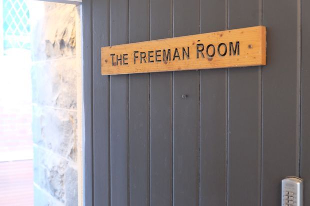 Close-up of a dark grey vertical-panelled wall with a rustic timber sign reading “THE FREEMAN ROOM.” A bright glazed opening and stone-textured surface are visible at the left edge, with a keypad or access device at the lower right. The image focuses on an entrance identification sign and building wall detailing.