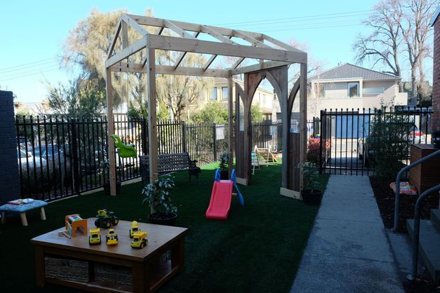 A fenced outdoor courtyard or backyard play area with artificial turf, children’s play equipment, and a timber-framed pergola structure. The pergola has an open pitched roof frame and decorative dark timber arched panels. A concrete walkway runs alongside the play space, with neighbouring homes and mature trees visible beyond the black metal fencing.