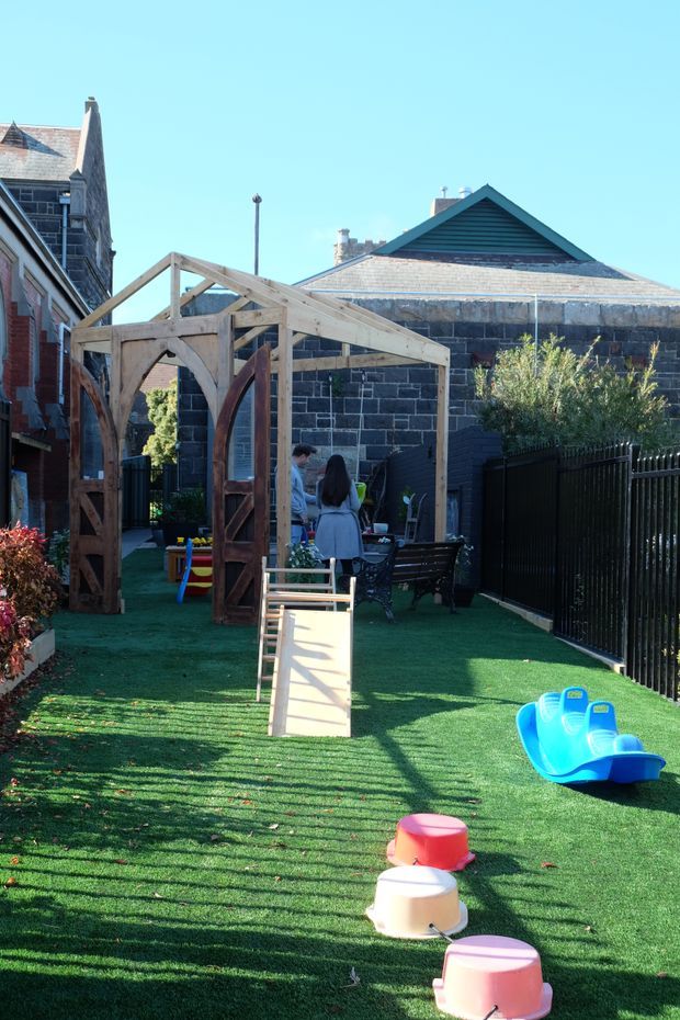 A narrow urban outdoor courtyard or play area with artificial grass, enclosed by dark fencing and historic masonry buildings. A timber-framed structure with arched gate openings occupies the centre, with outdoor seating, children's play equipment, and small planted areas. The scene is brightly lit under a clear blue sky.