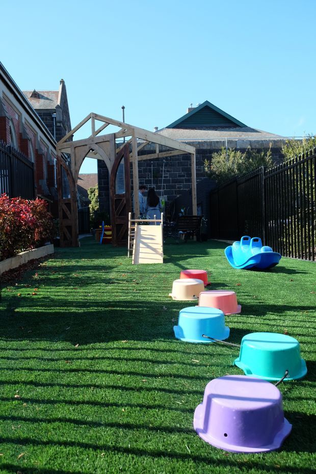 A sunny outdoor play area with artificial grass, colourful children's stepping pods, a blue rocker, and a timber play structure with a slide. The space is enclosed by dark fencing and bordered by brick and dark masonry buildings, with pitched rooflines visible in the background.