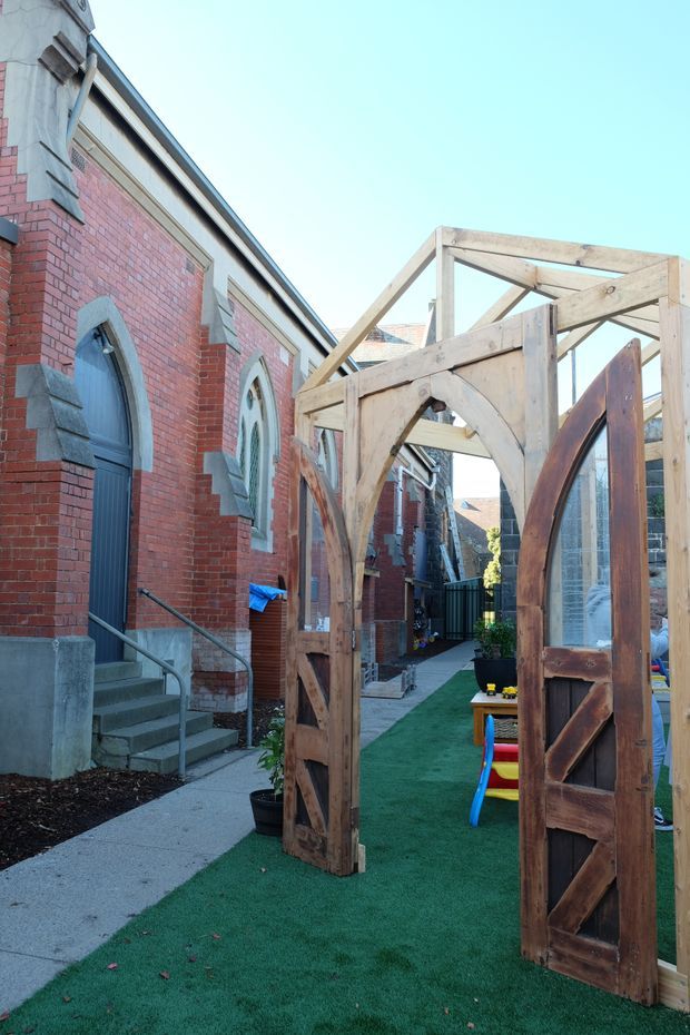 Exterior view of a red-brick heritage-style building beside a narrow landscaped courtyard or walkway. The facade features arched door and window openings with contrasting grey stone surrounds and detailing. In the foreground is a rustic timber-framed gateway or pergola structure with large wooden doors, opening onto artificial grass and a small play area.