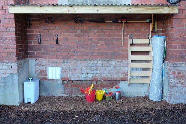 A rustic outdoor storage and play area against an exposed red-brick wall. A long timber shelf with hooks holds gardening tools above a small wooden ladder-style rack, while colourful children's beach or gardening toys sit on a bark-covered ground surface. A rolled patterned mat leans against the wall. The space is utilitarian and weathered, with corrugated roofing visible above.