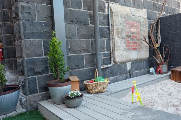 A small landscaped courtyard or outdoor play area beside a dark masonry wall. The scene features grey paved decking, potted plants, a wicker basket, exposed pipes, a weathered rectangular wall plaque, and children's beach toys in a light sandy section. The space has a utilitarian, historic-industrial character with subdued grey tones and natural plant accents.