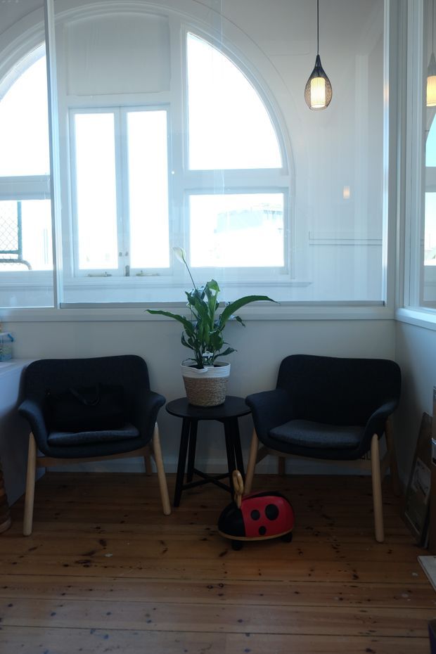 A compact, bright sitting area with two dark upholstered armchairs positioned around a small black side table holding a potted plant. The room has white painted walls with decorative arched window detailing, abundant glazing, pendant lights, and warm natural timber floorboards. A colourful children's ride-on toy is visible in the foreground.