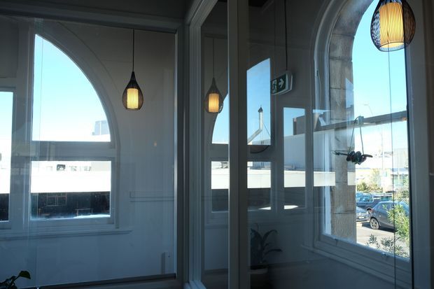 A bright enclosed entry or hallway with tall arched windows, dark-framed glazing, white paneled walls, and several warm pendant lights. Reflections in the glazing show the repeated arches and interior lighting. Urban buildings and a parked car are visible outside.