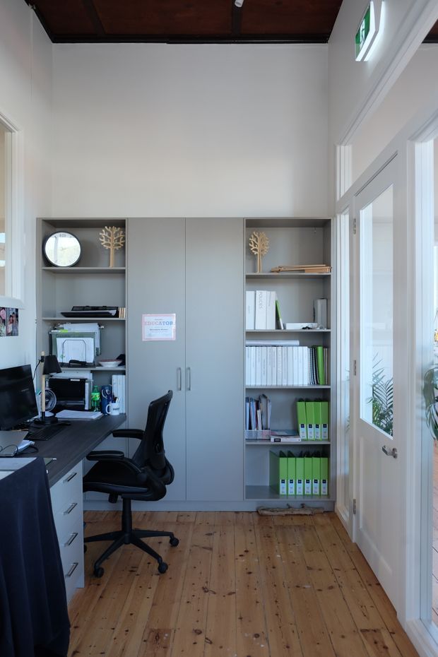A compact home office with a built-in grey storage wall featuring closed cupboards and open shelving filled with books, folders and office supplies. A dark desk with computer equipment and an ergonomic office chair occupies the left side. The room has warm timber floorboards, white walls and trim, glazed French-style doors on the right, and a dark timber-lined ceiling.