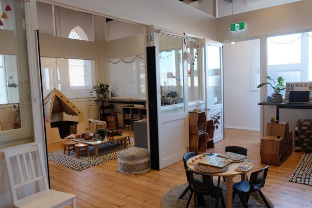 Bright, open-plan interior of a childcare or family play space with timber flooring, glazed internal partitions, white-painted walls and joinery, and multiple activity areas. A small children's table and chairs occupy the foreground, while a play tent, low shelving, toys, and soft furnishings are visible in the adjoining room. High windows and glazed doors bring in abundant natural light.