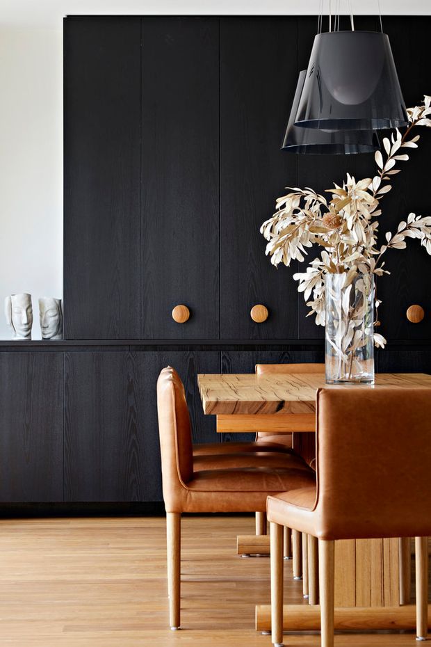 A refined dining area with a dark timber-panelled built-in wall and cabinetry, warm oak dining table and timber flooring, and caramel leather dining chairs. A large black pendant light hangs above a glass vase filled with dried pale foliage, creating a strong contrast against the dark joinery.