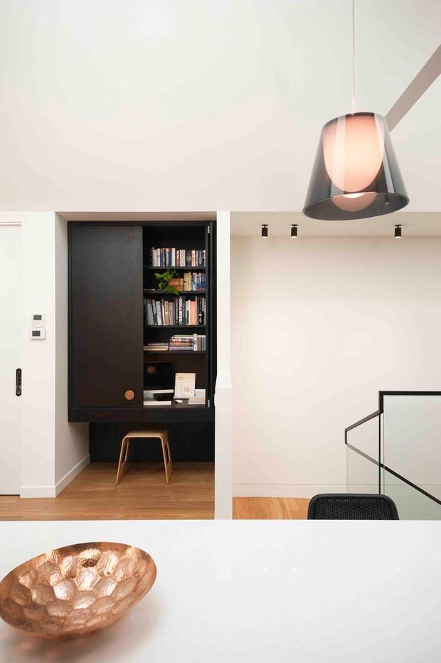 A bright contemporary interior showing a compact built-in black study nook with open bookshelves, a desk surface and a small timber stool. The nook sits beside a stairwell with frameless glass and dark metal edging. Warm timber flooring, white walls, recessed spotlights, and a large black pendant light define the space. A hammered copper bowl is visible on a white foreground surface.