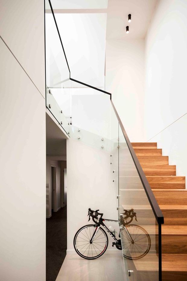 A bright, minimalist interior stairway with a sculptural open staircase ascending along the right side. The stair treads are warm natural timber, paired with clear glass balustrades and slim black handrails. White walls and a high ceiling create an airy vertical void, with a bicycle positioned beneath the stairs and a darker hallway visible beyond.
