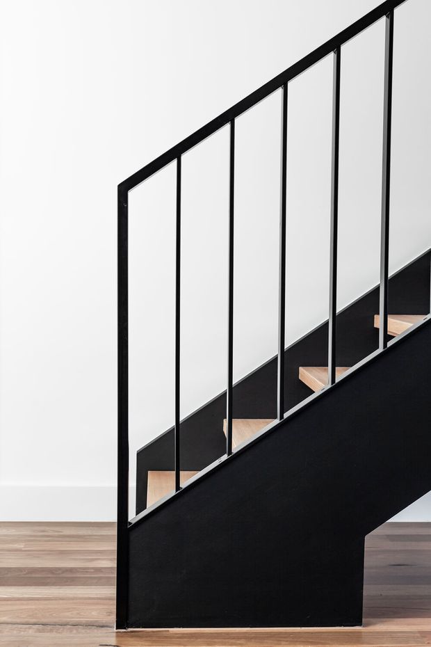 Minimalist interior view of a straight staircase with a solid black side stringer and matching black metal balustrade featuring slender vertical rods. Light timber stair treads contrast against white walls and a timber floor. The composition is graphic and contemporary, with clean lines and strong black-and-white contrast.