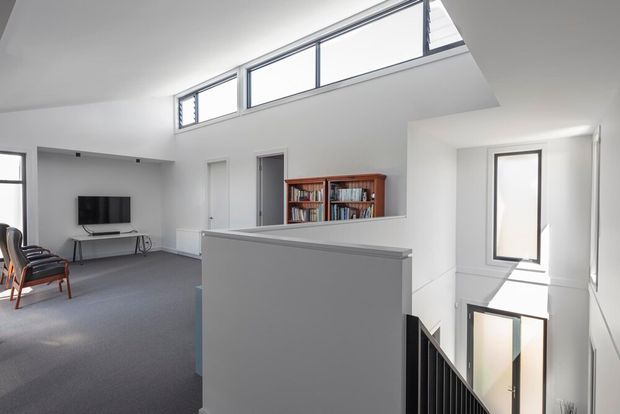 Bright contemporary multi-level interior viewed from a stair landing, with a double-height void, white walls, high clerestory glazing, grey carpet, black stair balustrade, and a small living area with television and timber-framed lounge chairs. A timber bookshelf sits along the upper-level wall, while strong daylight enters through the high windows and adjacent glazing.