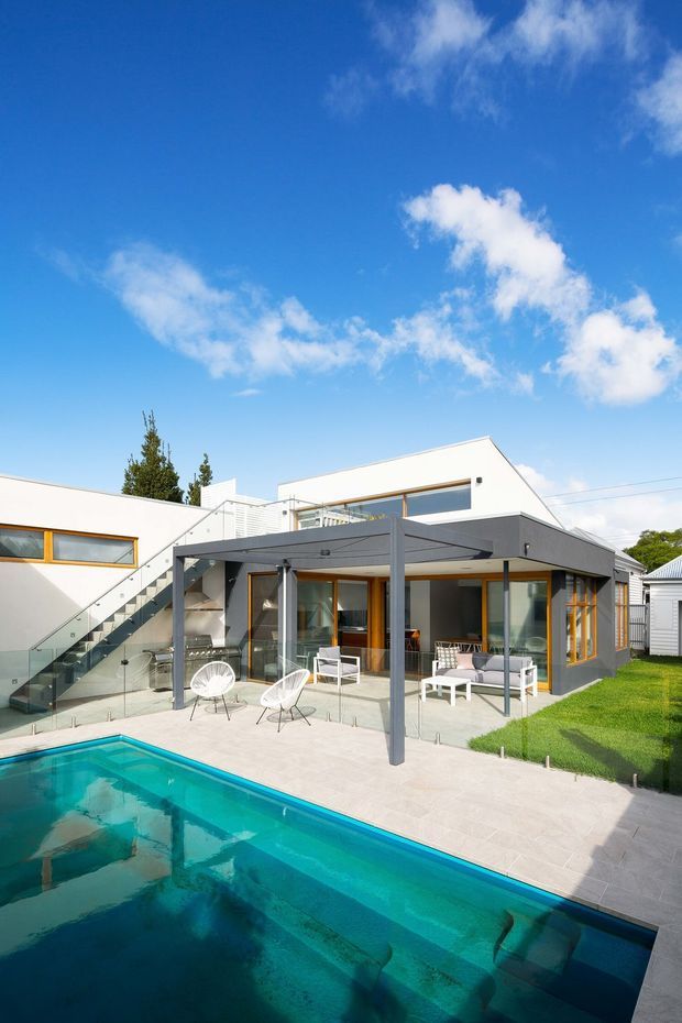 Bright exterior photograph of a contemporary multi-level home with white rendered walls, charcoal flat-roofed outdoor entertaining structure, extensive glazing with warm timber frames, an external glass-balustraded stair, paved pool terrace, and a swimming pool in the foreground. The landscaped backyard and clear sky reinforce the indoor-outdoor living focus.