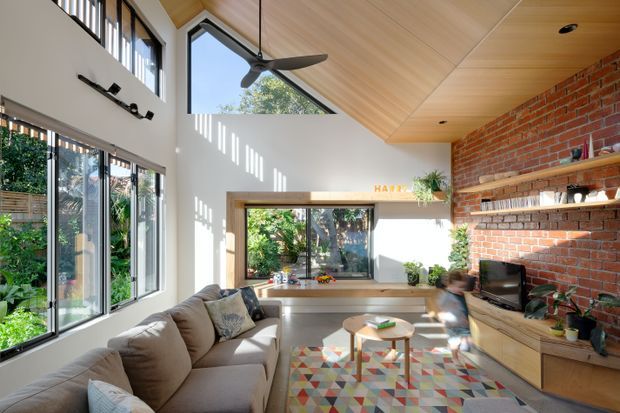 Bright contemporary living room with a vaulted timber-lined ceiling, exposed red-brick feature wall, extensive glazing, built-in timber window seating and cabinetry, a sectional sofa, and a colourful geometric rug. Sunlight creates strong linear shadows across the white walls, while open shelving and indoor plants add warmth and character.