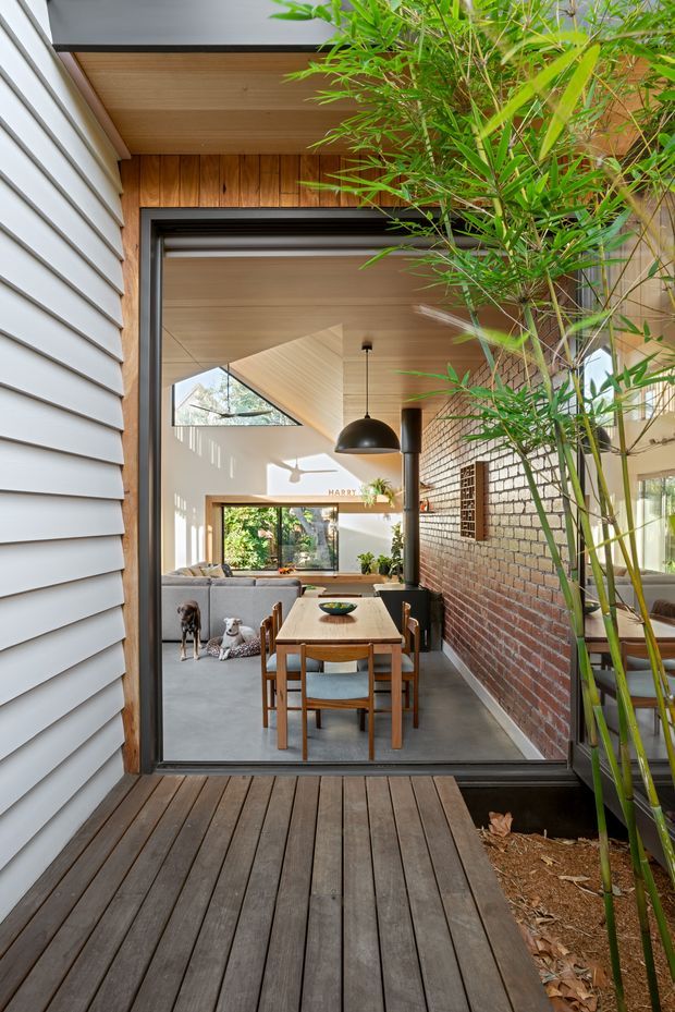 View from a timber deck through a large dark-framed opening into a bright open-plan dining and living area. The interior features a timber dining table and chairs, grey sofa, polished concrete floor, exposed red brick wall, timber-lined ceiling, pendant lighting, and extensive glazing. Grey horizontal weatherboard cladding frames the exterior, with bamboo planting beside the deck.