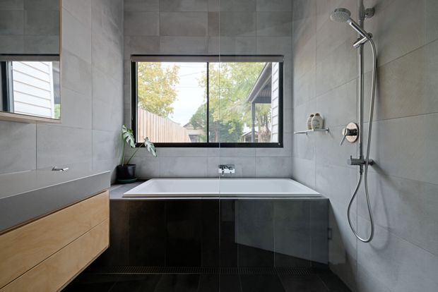 Modern bathroom with a freestanding-style rectangular bathtub set beneath a large window, a wall-mounted rainfall shower, floating timber vanity with a grey benchtop, and large-format grey wall tiles. Dark glossy tile panels surround the bath and continue across the floor. The room has a clean, restrained contemporary fit-out with natural light and views to neighbouring landscaping through the window.