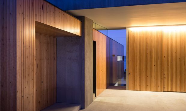 A contemporary architectural interior or entry passage with extensive vertical timber wall panelling, a large flush timber door or concealed storage wall, exposed concrete elements, and a pale smooth floor. Warm linear lighting highlights the timber surfaces, while glazing at the rear reveals a cool blue exterior evening light. The composition is minimalist and corridor-like, with strong geometric planes and built-in joinery.