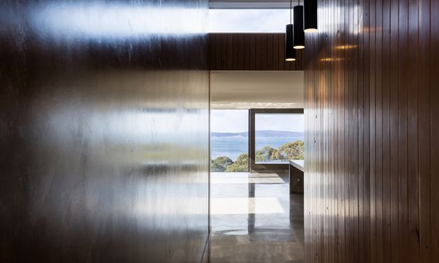 A dark, narrow interior passage framed by reflective dark wall panels on the left and vertical timber wall panelling on the right. Black pendant lights hang from the ceiling, while a glazed opening at the far end leads toward a bright outdoor terrace with expansive water and landscape views. The polished concrete floor and strong contrast between the moody corridor and sunlit exterior create a dramatic, contemporary architectural composition.