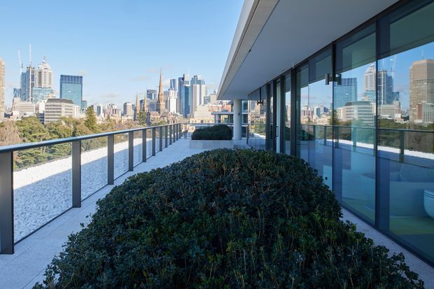 A wide elevated terrace or balcony outside a modern building, with dark-framed floor-to-ceiling glazing, a deep projecting roof slab, glass balustrades, pale paving, and integrated dense green shrub planting. A contemporary city skyline is visible beyond the terrace.
