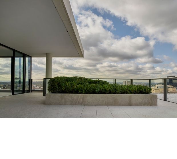 A contemporary rooftop terrace or balcony with a broad cantilevered roof, full-height glazed doors, pale tiled flooring, and a long integrated stone planter filled with clipped greenery. A slim metal balustrade frames expansive urban views beneath a partly cloudy sky.