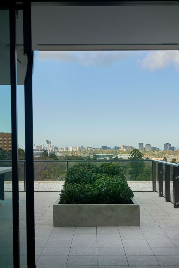 View from an interior through full-height glazing onto a tiled terrace or balcony with a large rectangular planter and manicured shrub. A glass balustrade frames a broad urban skyline and landscaped park beyond. The scene is clean and contemporary, with a partially lowered roller blind overhead and dark window framing.