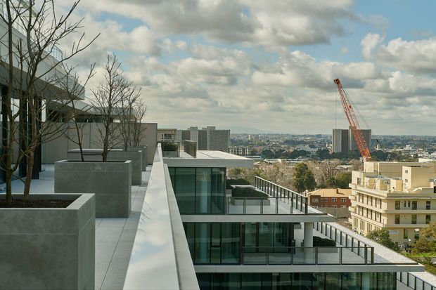 View from a contemporary multi-level building terrace or rooftop, with large concrete planters, minimalist paving, glass balustrades, and layered flat-roofed forms. The scene overlooks a dense urban neighbourhood with mid-rise buildings and a construction crane under a partly cloudy sky.