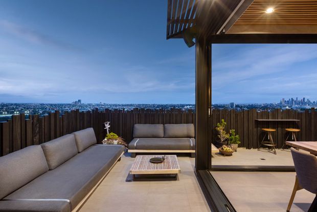 A contemporary rooftop terrace at dusk with a covered outdoor lounge, large sectional sofa, coffee table, and additional seating arranged on pale tiled flooring. A dark vertical timber privacy screen frames the terrace, while a slatted timber ceiling with integrated downlighting creates a warm sheltered atmosphere. The open side overlooks a dense illuminated city skyline.