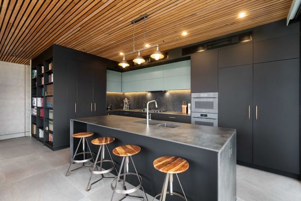 Contemporary open-plan kitchen with a large dark island and four timber-topped stools, matte dark cabinetry, integrated appliances, a metallic stone-look benchtop, open shelving, and a striking slatted timber ceiling. The space has a refined industrial character with concrete-look flooring and exposed ductwork or ventilation details.