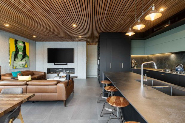 Open-plan contemporary kitchen and living area with a large stone island and integrated sink, dark cabinetry, timber slatted ceiling, concrete-look wall panels and flooring, pendant lighting, and a brown leather sectional surrounding a media wall.