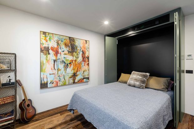Compact contemporary bedroom with a wall-integrated fold-down bed or Murphy bed, framed by dark cabinetry and open shelving. The room has white walls, warm timber flooring, neutral bedding, an abstract artwork, and a guitar with shelving along the side wall.