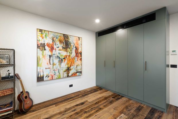 A compact modern room with full-height muted grey built-in cabinetry along one wall, integrated vertical handles, and a dark horizontal overhead ventilation or display band. The room has white walls and ceiling, warm distressed timber flooring, a colourful abstract artwork, and a guitar with open shelving along the opposite wall. Recessed ceiling lighting and minimal detailing create a clean contemporary fit-out.